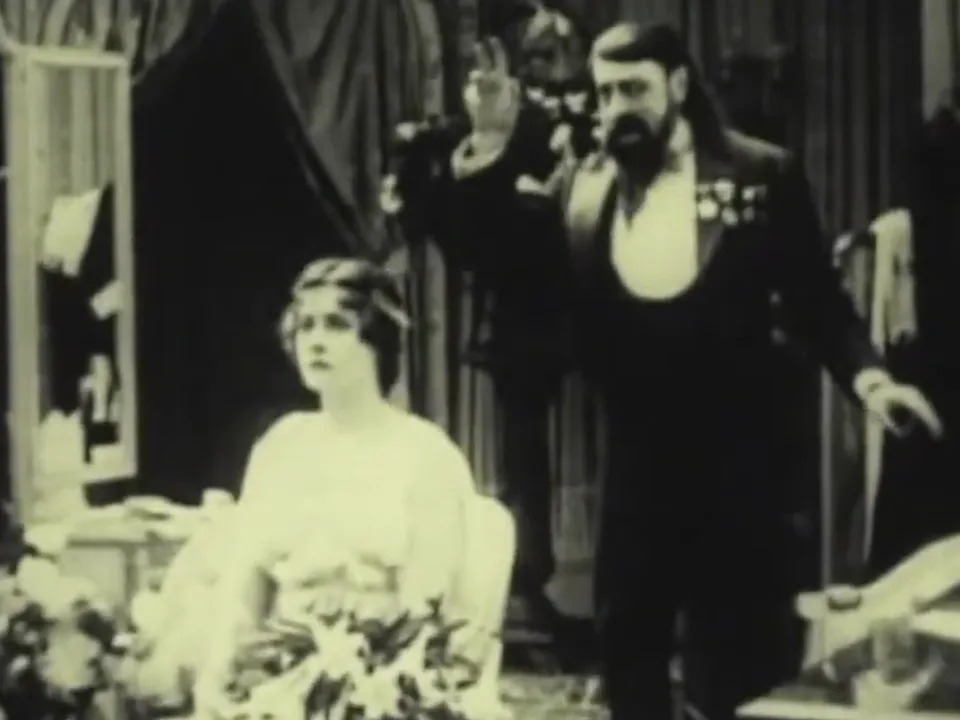 in a scene, Trilby sits hypnotized to stillness in a wedding dress, while Svengali stands in a tux behind her, talking while gesturing wildly
