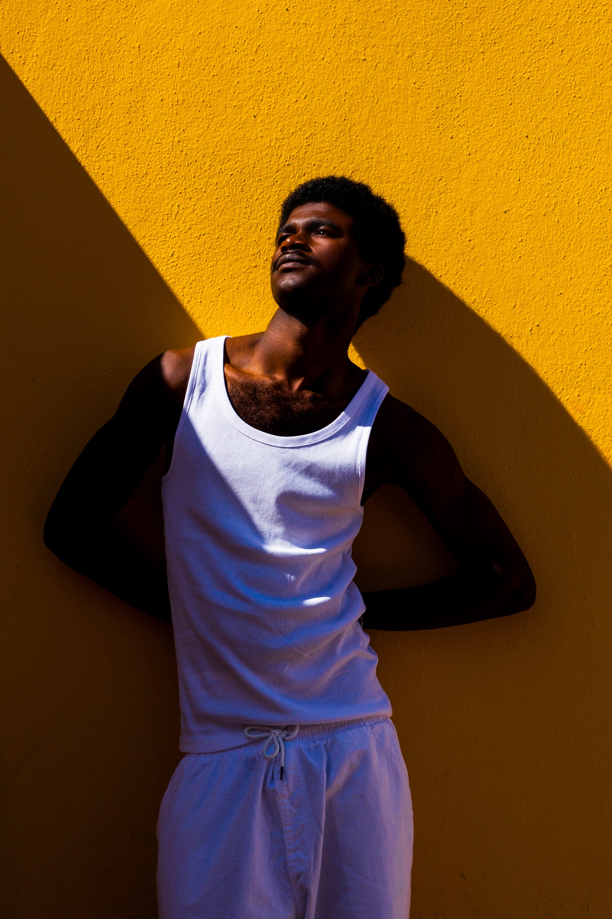 a photograph by Yannis Guibinga of a Black man wearing a white sleeveless shirt, standing in front of a bright yellow wall