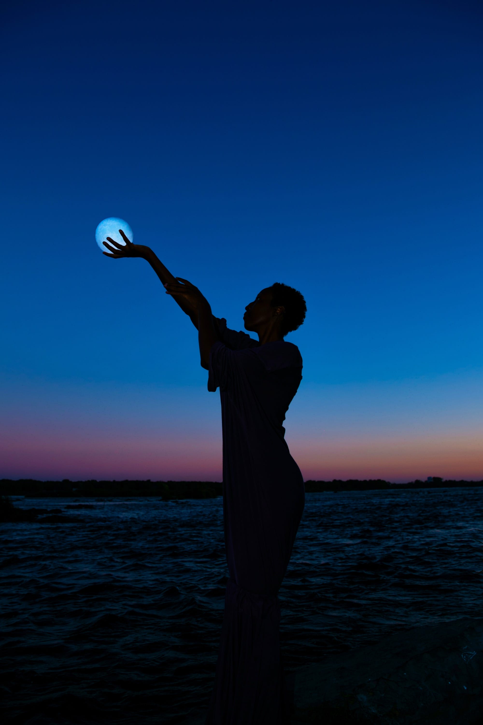 a photograph by Yannis Guibinga of a silhouetted figure holding their hand out to appear to hold the moon