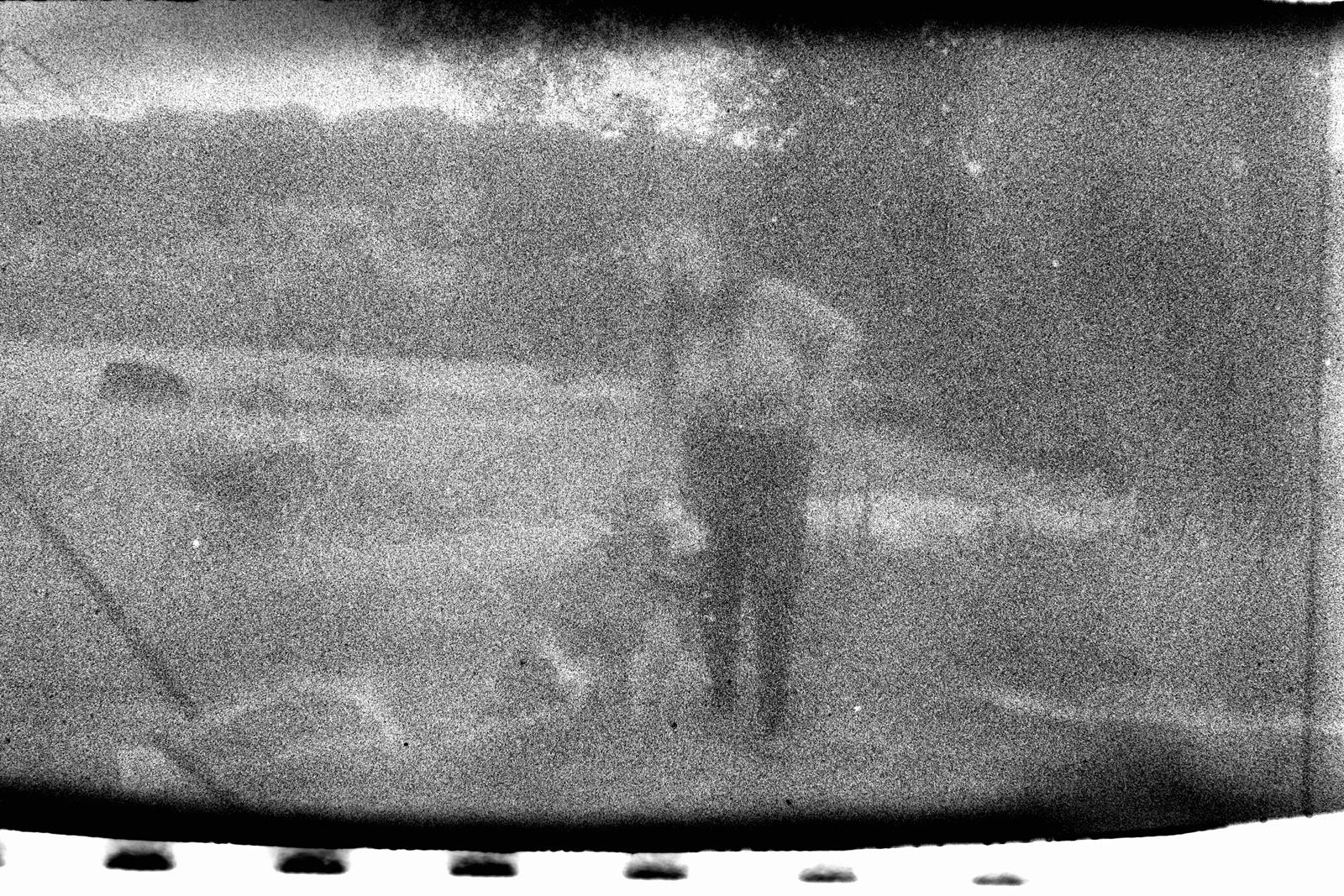 A grainy, black-and-white photo shows a person standing outdoors with a dog on a leash. The background is blurry, with trees and open land visible. The image appears to be damaged or taken with old film.