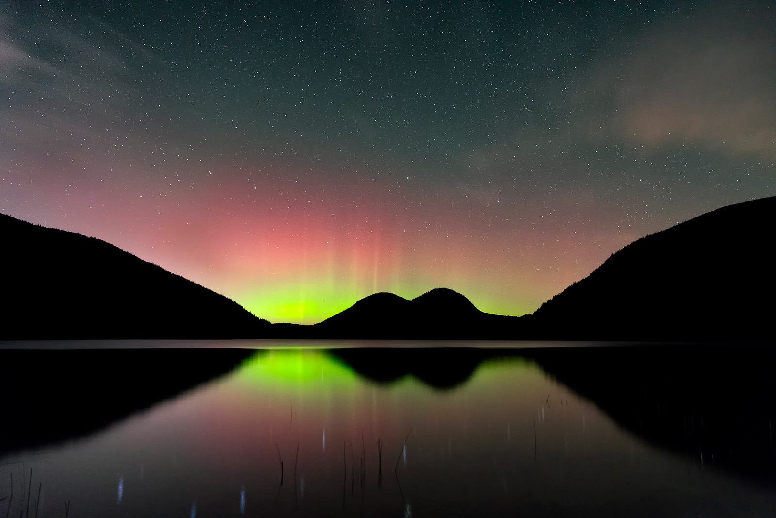 A serene lake reflects silhouetted hills under a night sky with colorful aurora lights in green, pink, and red hues, and countless stars twinkling above.