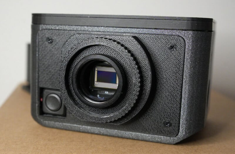 A close-up of a black digital camera with a textured body, prominent lens, and visible image sensor, placed on a brown surface against a light background.