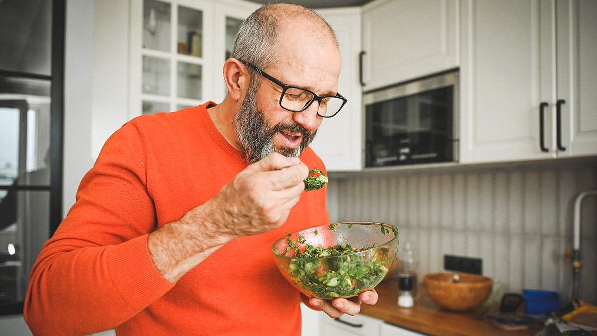 Older man wearing an orange sweater eating a fresh green salad from a glass bowl in a kitchen.