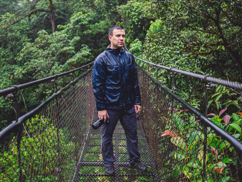 A man in a dark rain jacket stands on a metal suspension bridge in a lush, green forest, holding a camera in his right hand. The bridge is wet, and rain droplets are visible on his jacket.