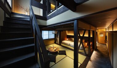 A wooden staircase and banister go up the left side of the photo, with a vase and flowers at the top of the stairs. A warmly lit, open plan area contains a small coffee table with futon-cushions for sitting and two beds towards the back of the space.