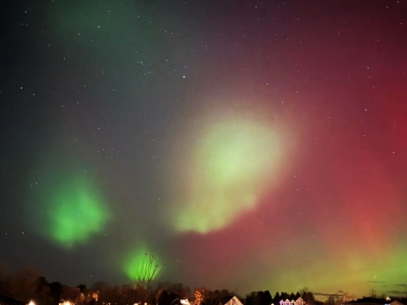 Reddish green aurora with bright green blobs in the foreground and the tops of houses below.