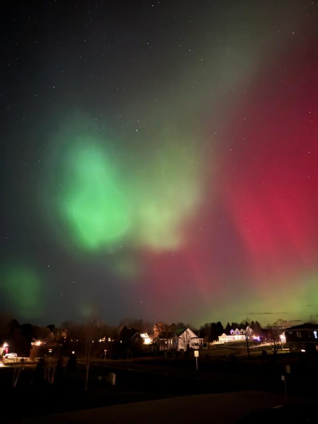Reddish green aurora with bright green blobs in the foreground and a few lit houses below.