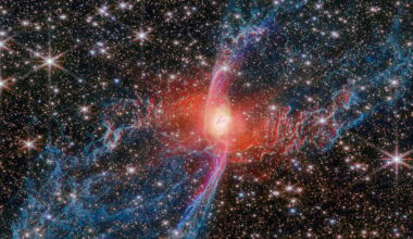 A vibrant nebula with a glowing red center and intricate blue and pink filaments stretches across a star-filled background in outer space. Many bright stars are scattered throughout the scene.