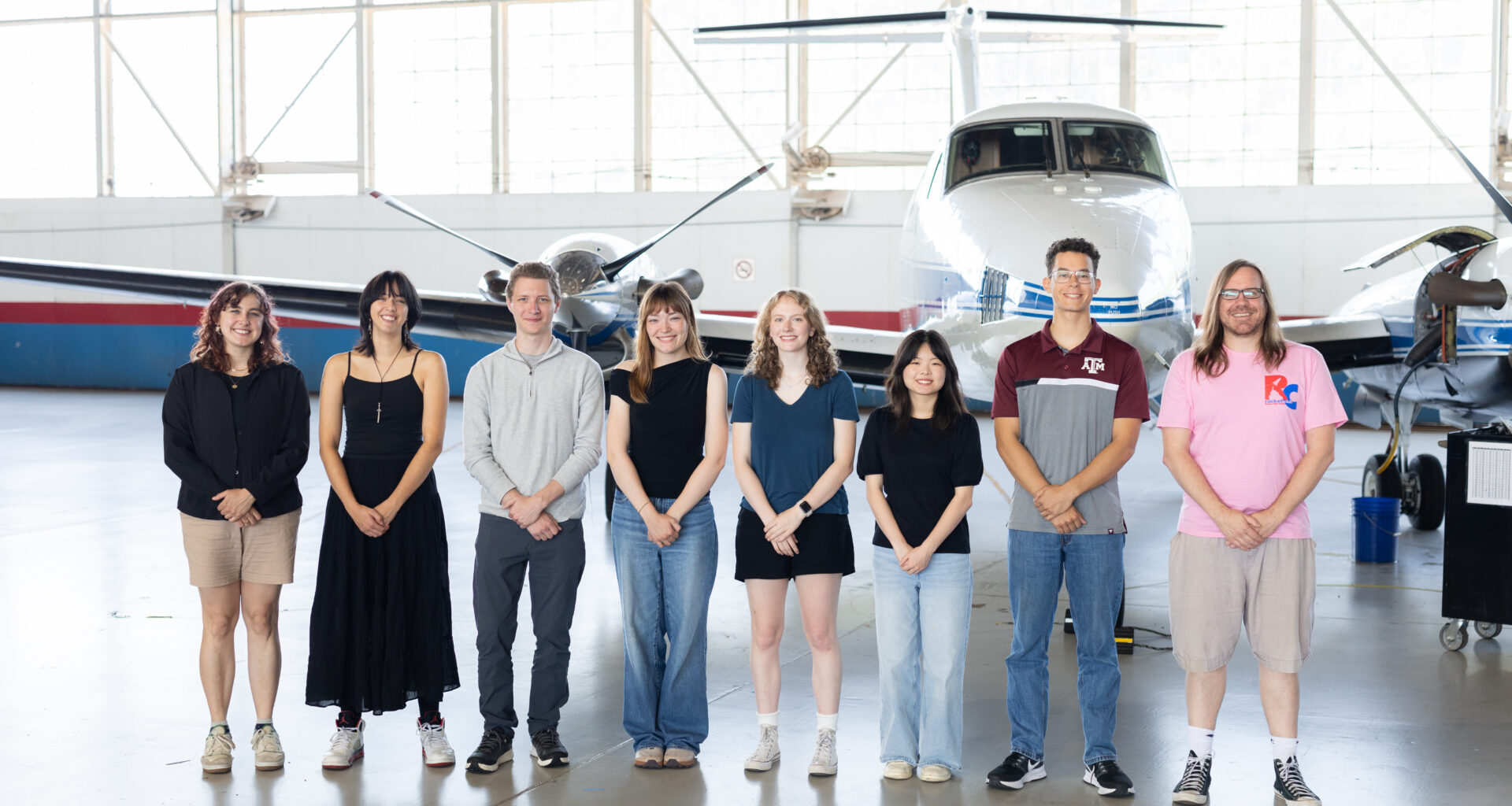 A group of eight people stand together inside a hangar with a somewhat shiny floor. In the background is a small white plane with a blue stripe, and large windows behind that.