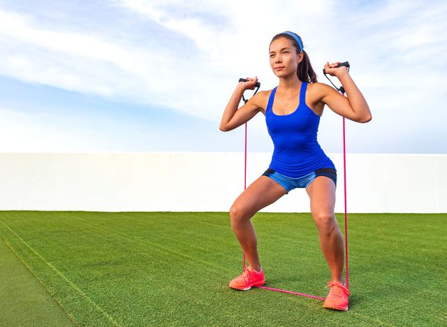 Exercise woman with fitness elastic band workout in outdoor gym . Asian girl doing resistance training outside on grass. Fit people lifestyle. Squat and shoulder press with elastics.