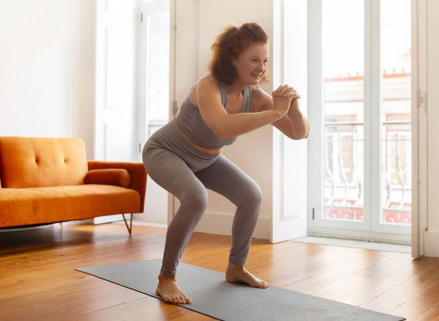 Smiling senior woman making squat exercise at home, active sporty elderly lady training on her yoga mat, smiling during fitness workout in her airy, well-lit living room, copy space