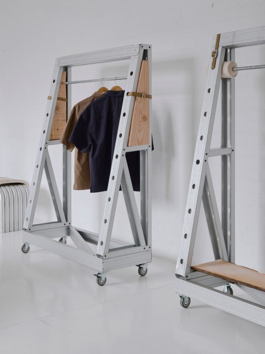 Minimalist clothing rack setup with industrial steel frames and wooden panels on caster wheels, displaying neutral-toned shirts on hangers in a white studio space.