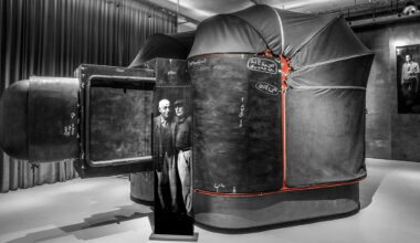 A large, dark, tent-like camera obscura with red accents sits in a gallery. An old black-and-white photograph of two men stands in front of it, and black curtains line the background wall.