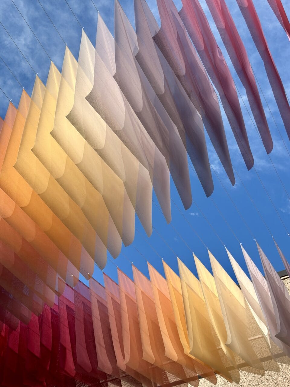 a detail of an art installation by Tomislav Topić of layers of colorful planes of fabric suspended above a street, between two buildings, in undulating rows so that it looks prismatic