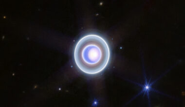 Uranus and its moons and rings glow blue, white, and teal in this infrared image. Uranus is seen nearly face-on, and moons are visible both within and outside of the ring system.