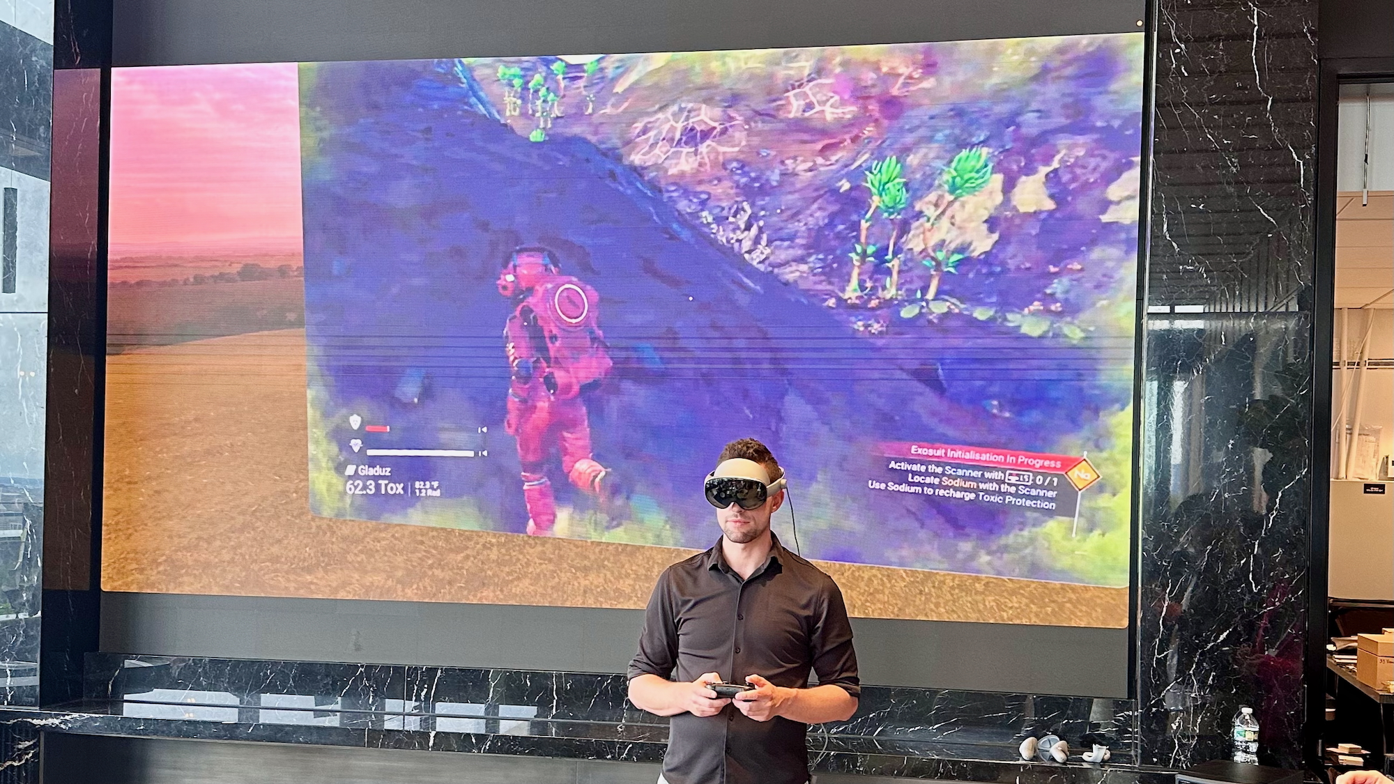 A photo of a Samsung engineer playing No Man's Sky on the Galaxy XR headset using a Bluetooth gamepad, with his headset feed visible on a screen behind him.