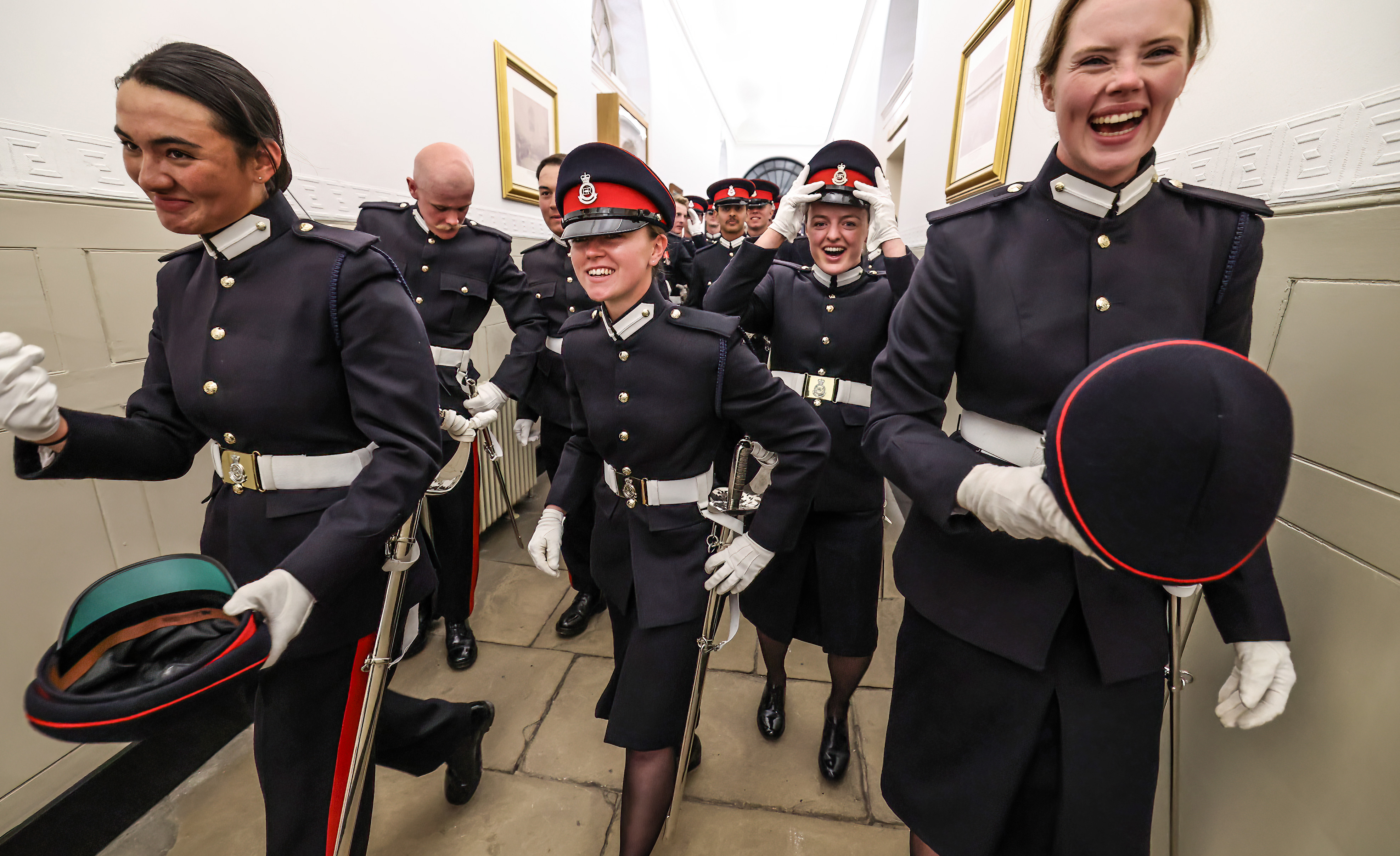 Smiling officer cadets in military uniforms parade through a corridor.