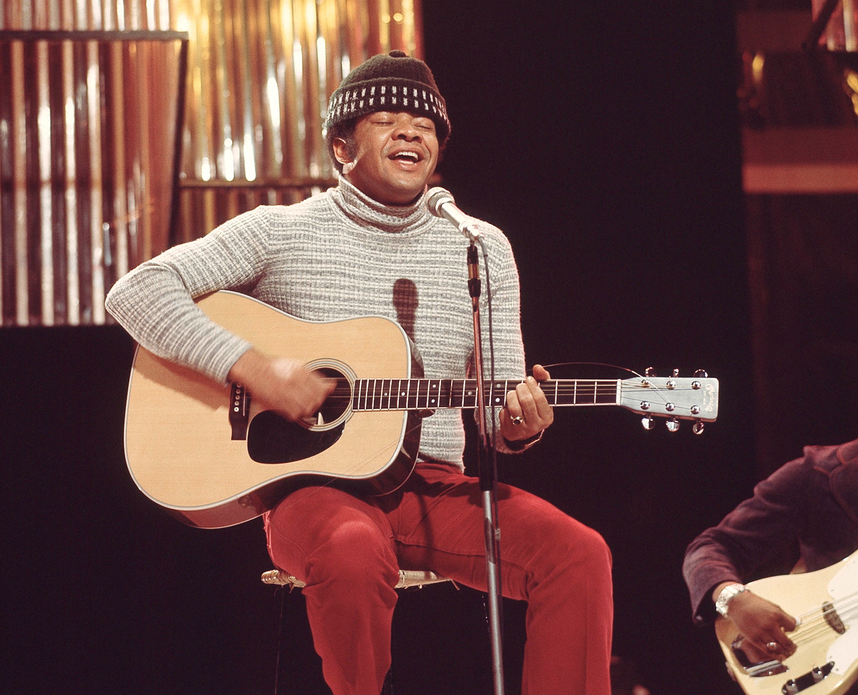 Bill Withers performing live on a UK TV show in London, playing an acoustic guitar and singing into a microphone.