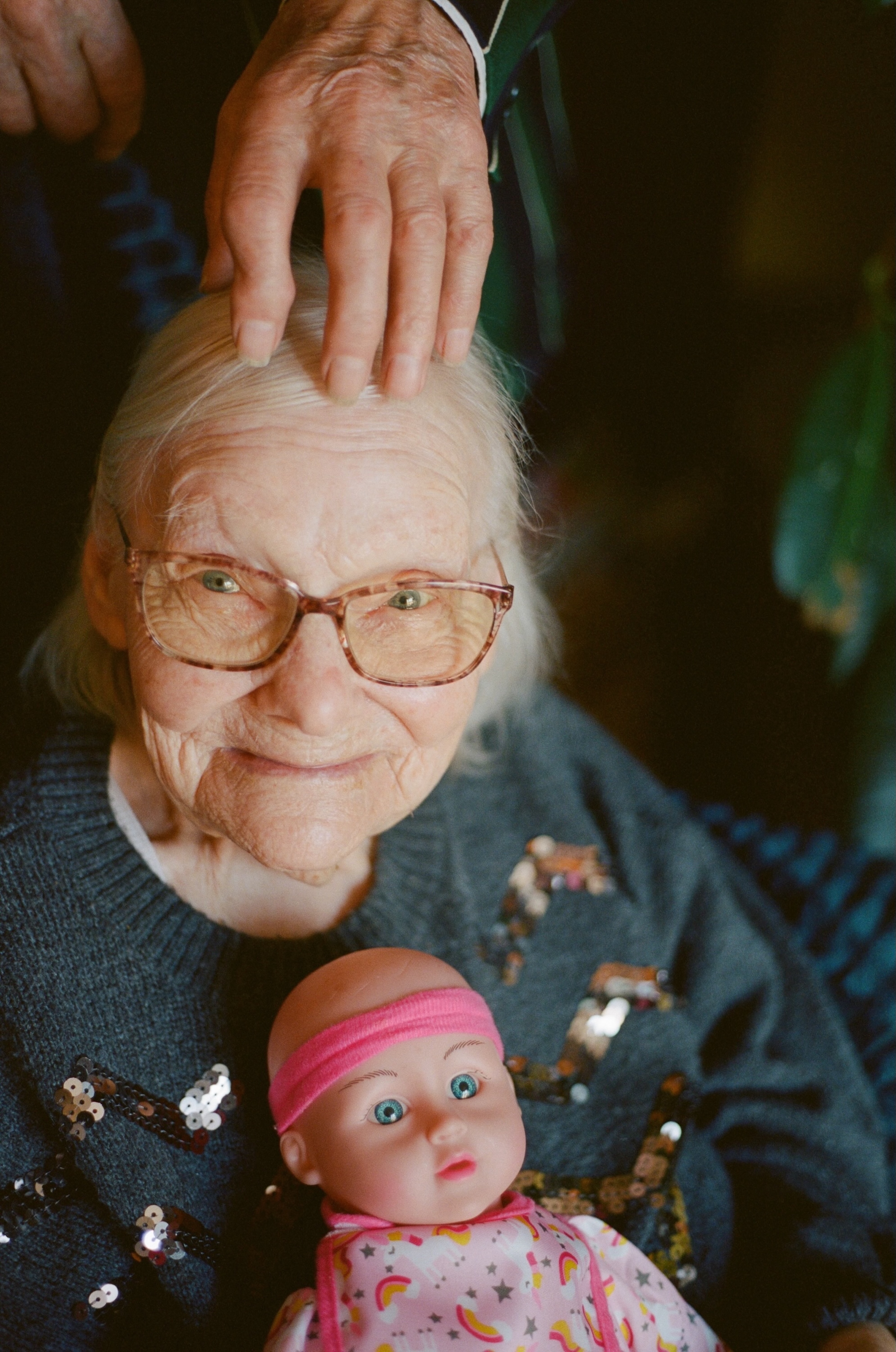Dorothy, a 95-year-old woman with dementia, smiles as Al places his hand on her head, while she holds a baby doll wearing a pink headband and a onesie with unicorns and stars.