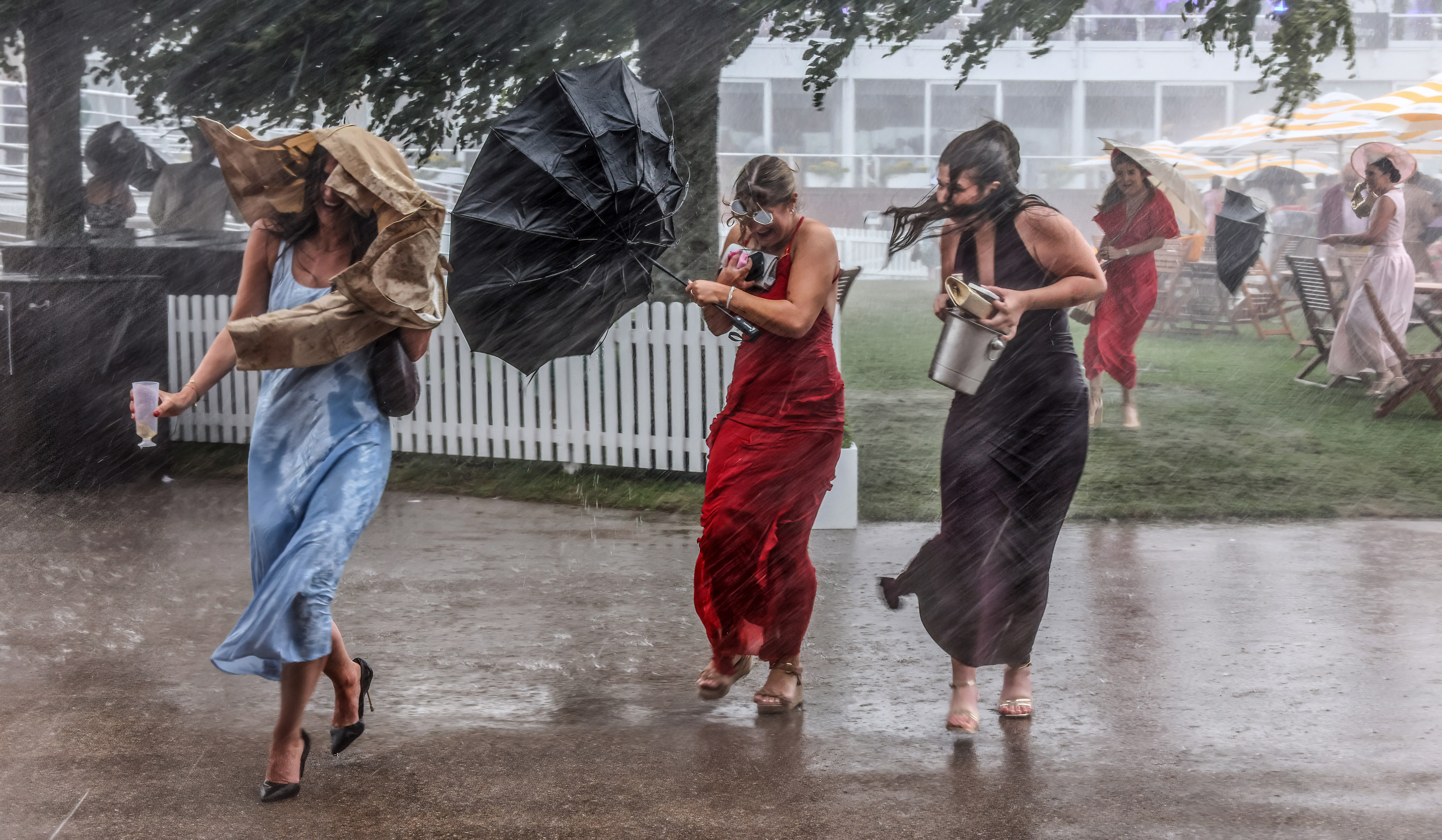 Three women run from a rainstorm, clutching drinks and an umbrella.