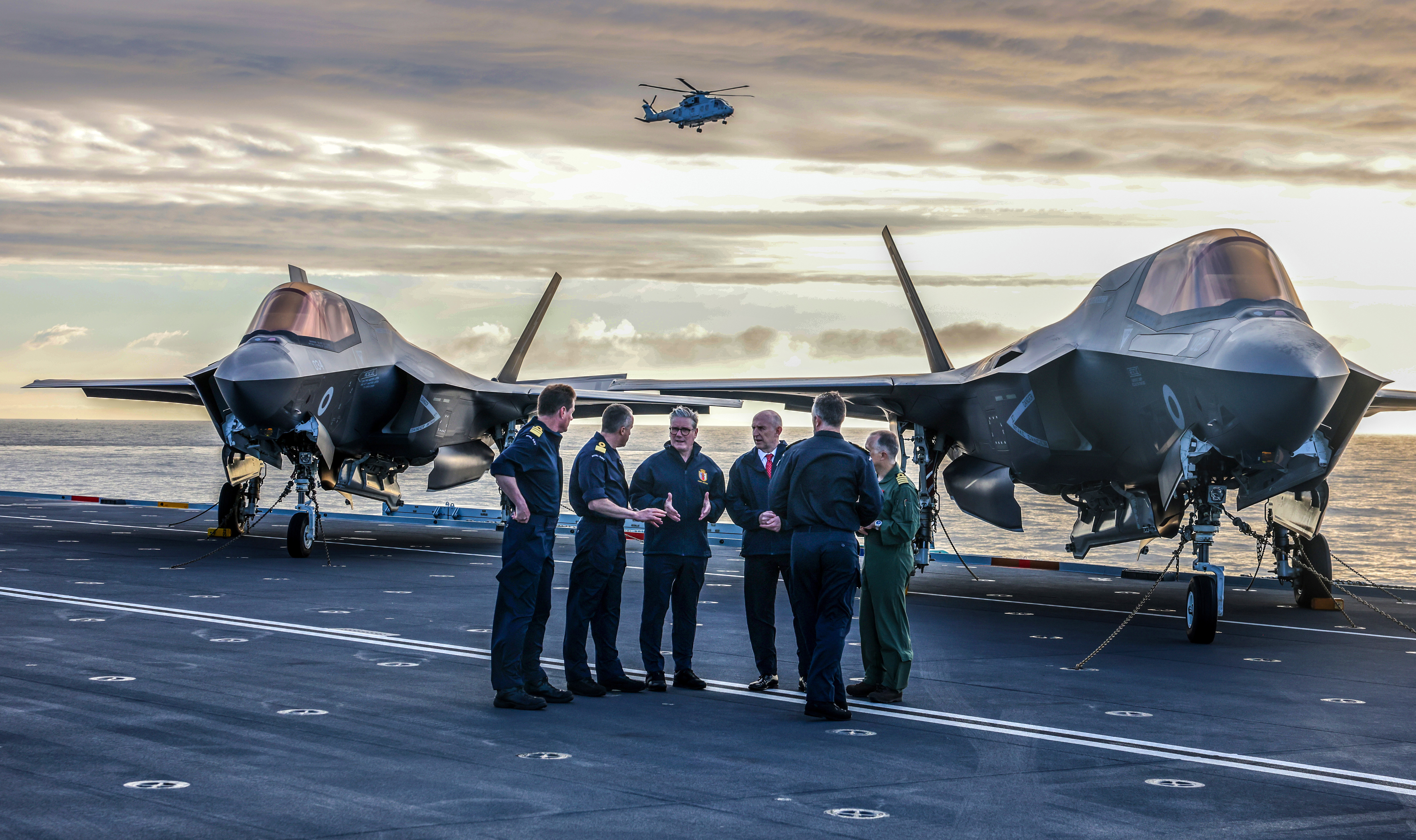 Prime Minister Keir Starmer and Defence Secretary John Healy talking to ship's officers in front of F-35 fighter jets on an aircraft carrier at dawn, with a helicopter overhead.