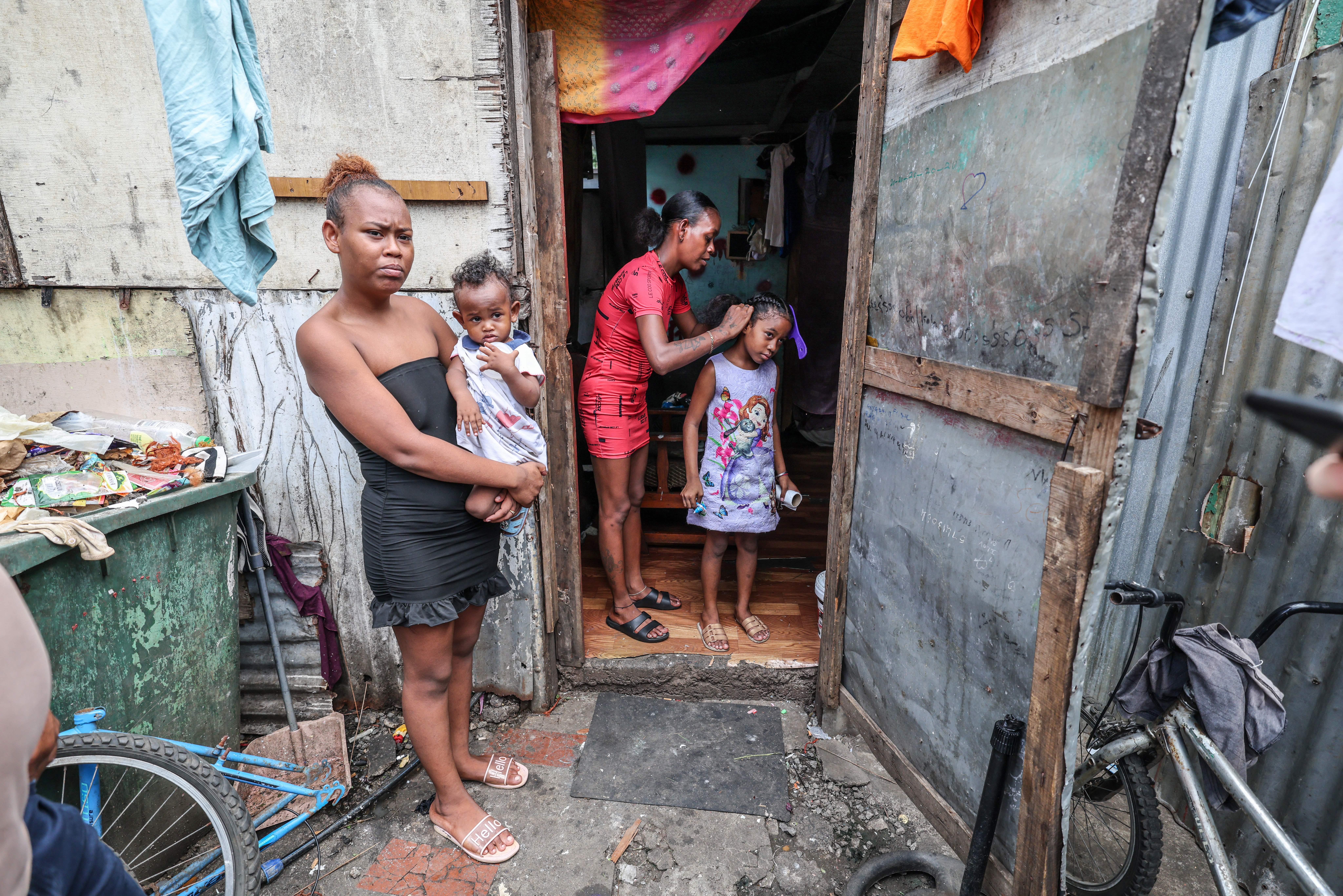 A woman holds a baby, while another woman braids a girl's hair in the doorway of a corrugated iron home.