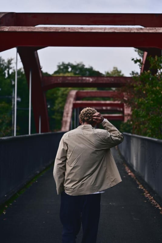 A person with short curly hair, seen from behind, walks alone on a bridge with red metal arches, surrounded by trees and overcast skies.