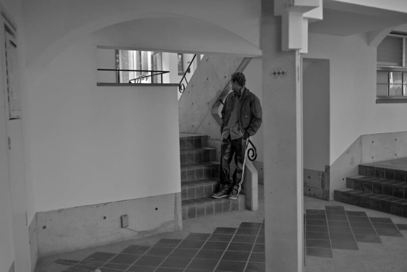 A person stands alone on an indoor staircase, leaning against the railing in a minimalist, modern building. The photo is in black and white, emphasizing shadows and architectural lines.