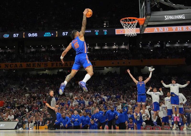A basketball player in a blue uniform leaps mid-air for a dunk during a game, with teammates and fans cheering in the background. The scoreboard shows a tied score of 57-57 and 9:56 left in the second half.