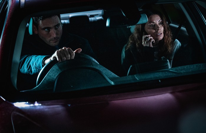 Kit drives a darkened car as Carla, in the passenger seat, uses her phone in a scene from Corriedale