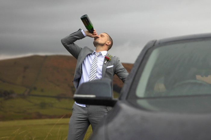 Carl Webster, stood in moorland, swigs from a bottle in a wedding suit next to a car in a scene from Coronation Street