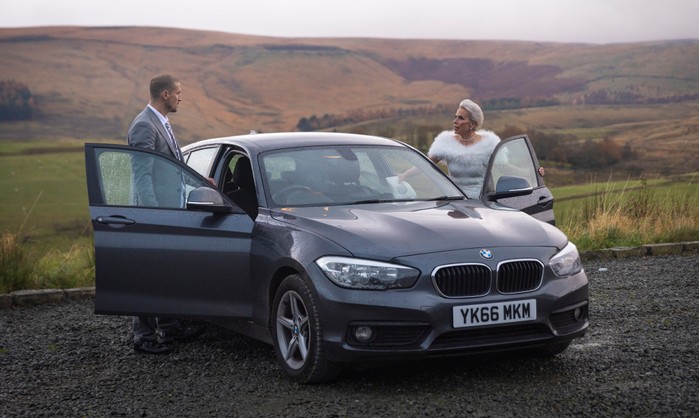 Debbie, in her wedding dress, stands opposite Carl, in a suit, as they are surrounded by hills and next to a car in a scene from Coronation Street