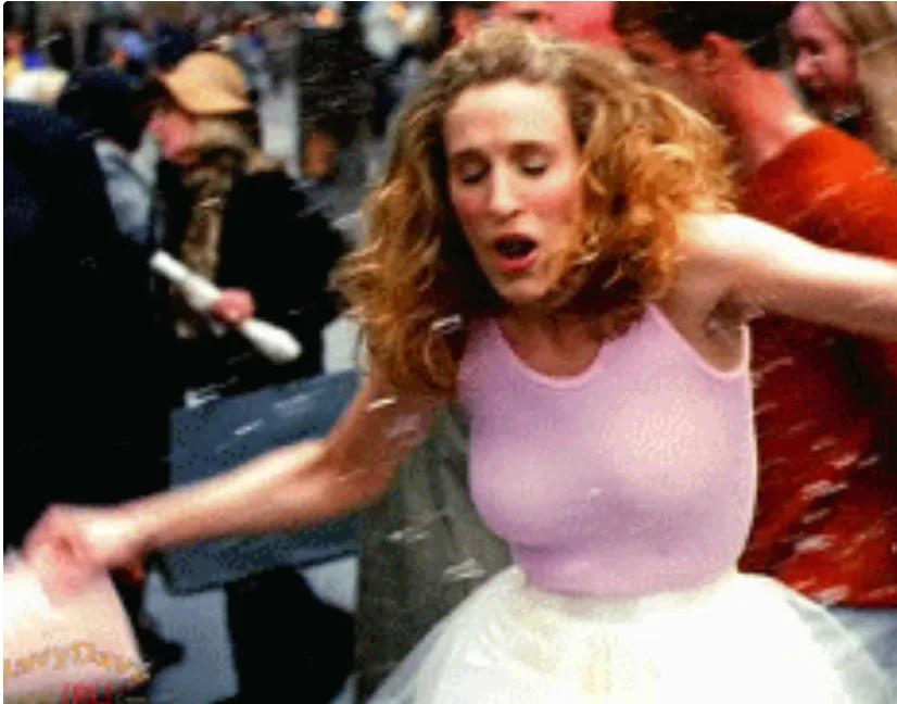 Person with curly hair in a sleeveless top and tulle skirt playfully interacts with floating confetti on a busy street