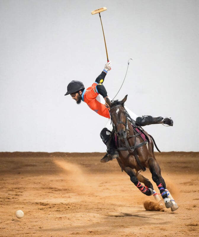 A polo player in an orange shirt leans dramatically off his horse while swinging his mallet toward a ball on the sandy ground during an intense match.