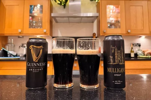A can of Guinness, with its contents poured into a pint glass, next to a can of Mulligan's, which has also been poured into a pint glass. The cans and glasses are on a kitchen work surface