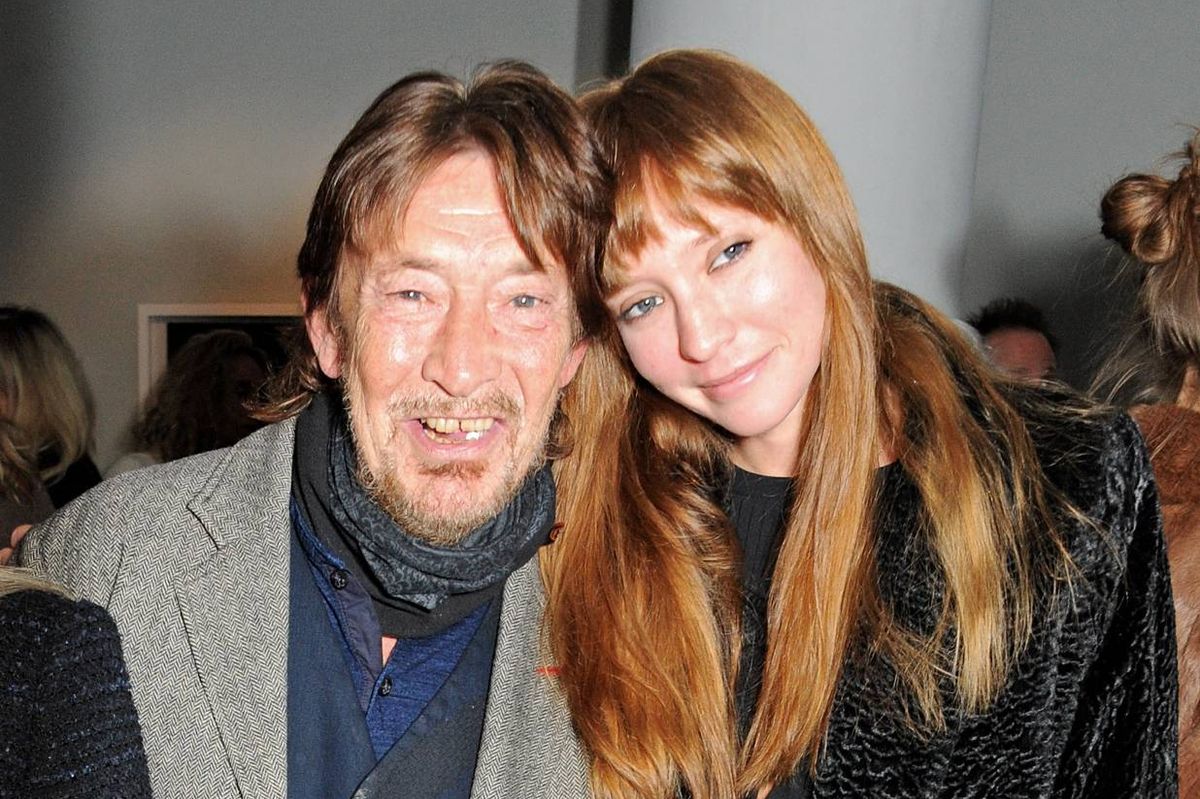 An image of two individuals standing close to each other, posing for a photograph with a backdrop of a room that appears to be an art gallery. The individual on the left is dressed in a blue shirt and a gray blazer, while the individual on the right is wearing a black top with long, flowing hair.