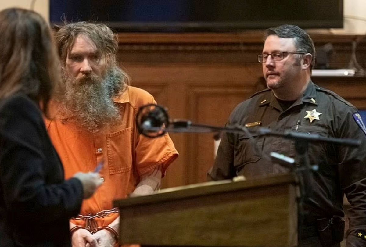 An individual in an orange prison uniform is being escorted by an officer in a courtroom setting. Another person in a dark jacket is present, engaged in an interaction with the individual in the orange uniform. A microphone is positioned on a podium in front of them.