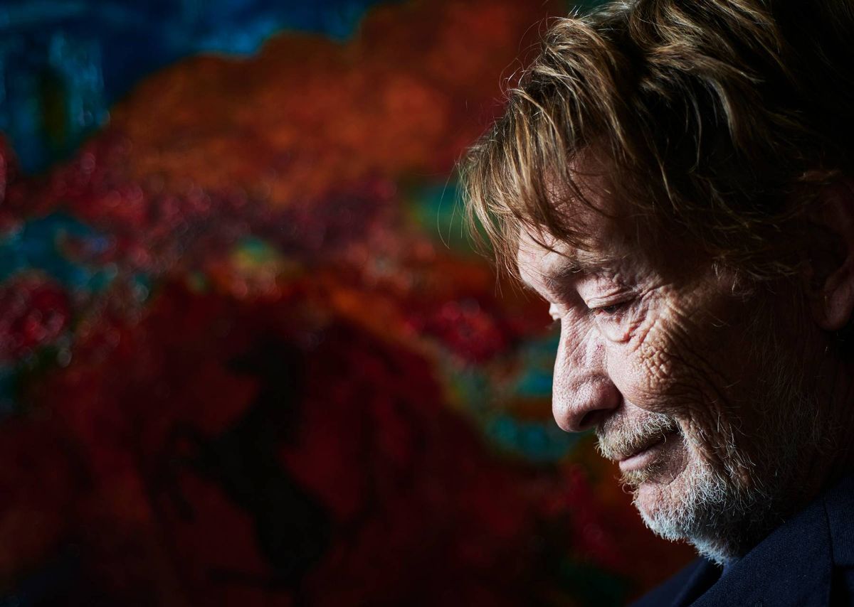 An individual with short hair, dressed in a dark-colored garment, is gazing contemplatively with eyes closed, positioned against a backdrop of vibrant red and green hues.