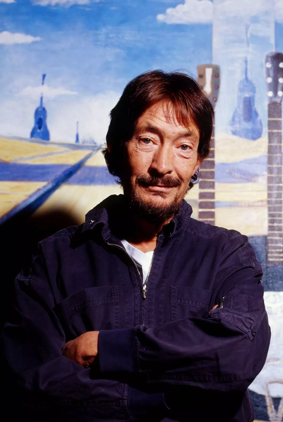 An individual with a mustache and a smiling face is standing in front of a backdrop that features a road with a blue and yellow hue and a tower.