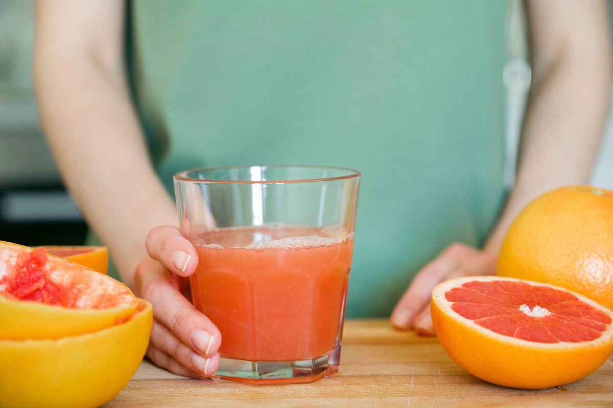 An individual holding a glass filled with a refreshing red beverage, standing in a kitchen setting with fresh citrus fruits nearby.