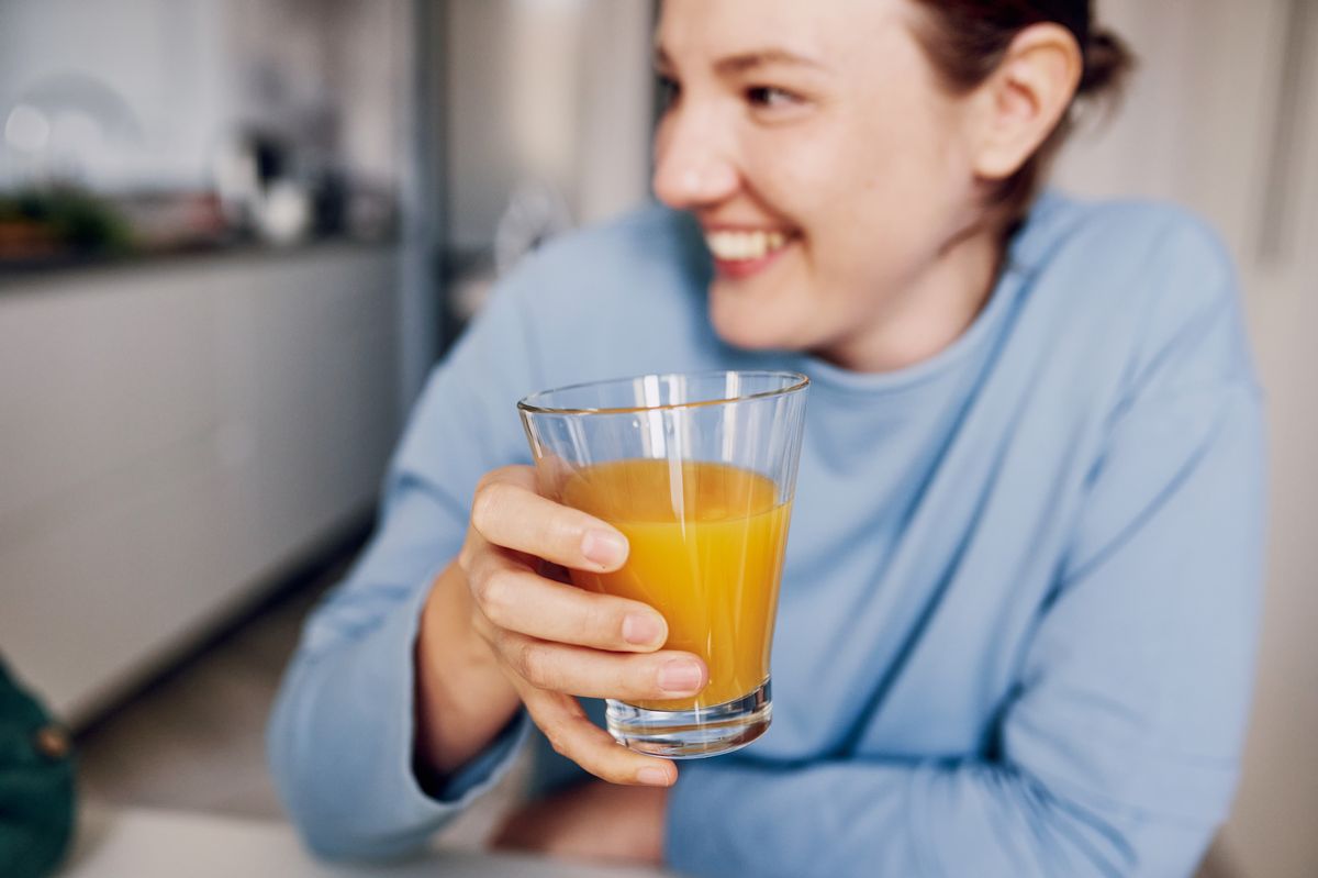 A cheerful woman in a light blue shirt holds a glass of orange juice, smiling and looking away