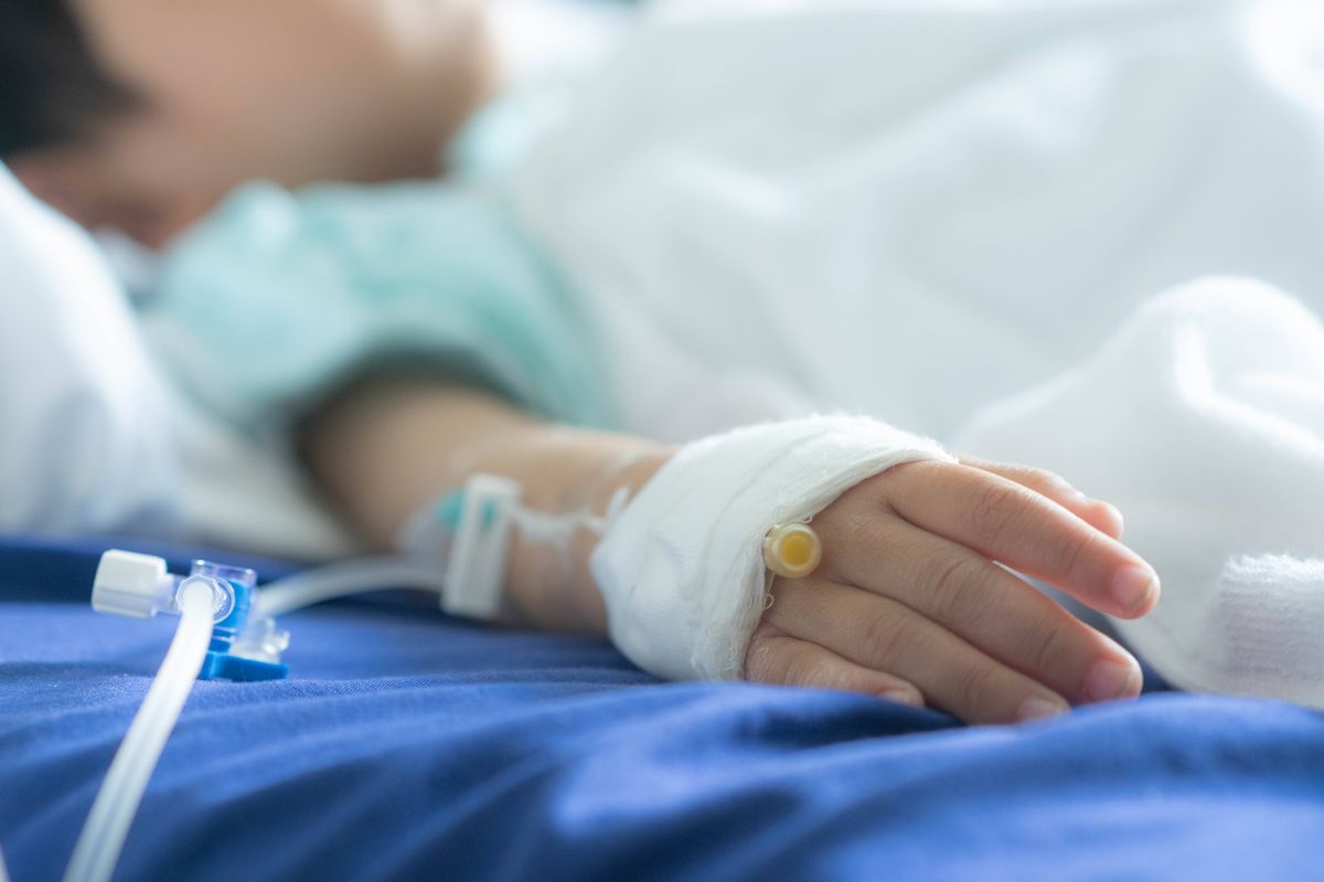 An individual in a hospital lies in bed with medical equipment connected to their arm, including an IV line and a bandage, while appearing to be in a state of rest.