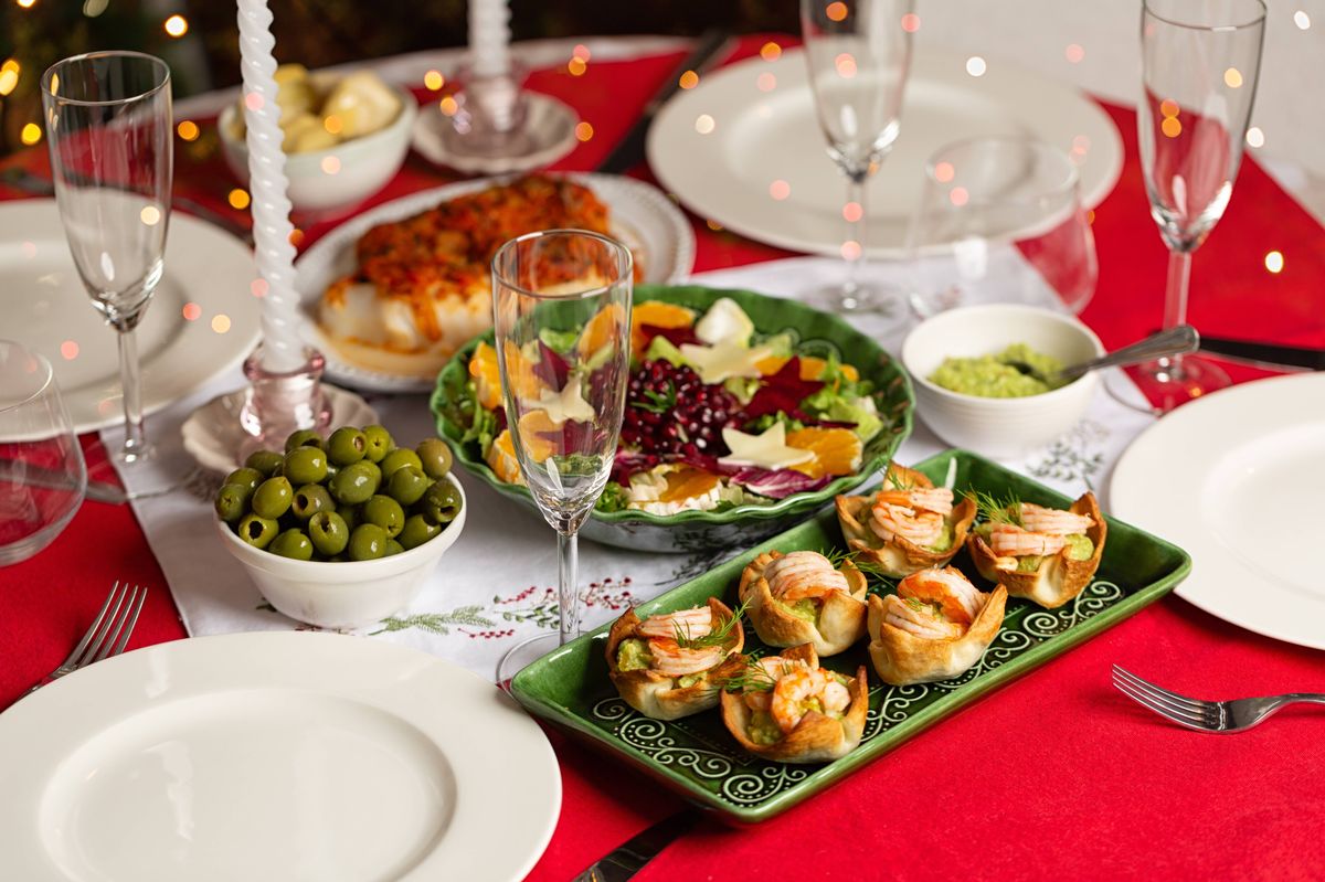 Christmas or New Year table with red tablecloth. Homemade food, canape, prawn and guacamole tartlets, winter salad, olives, greek fish, glasses.  Life style foto of dinner party.