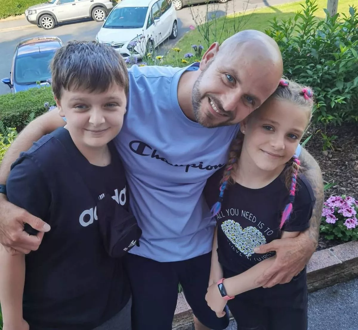 An adult male with a beard is standing with two children, one on each side, posing for a photograph in a garden with various plants and flowers. Behind them, a white car is parked on the side of the road.