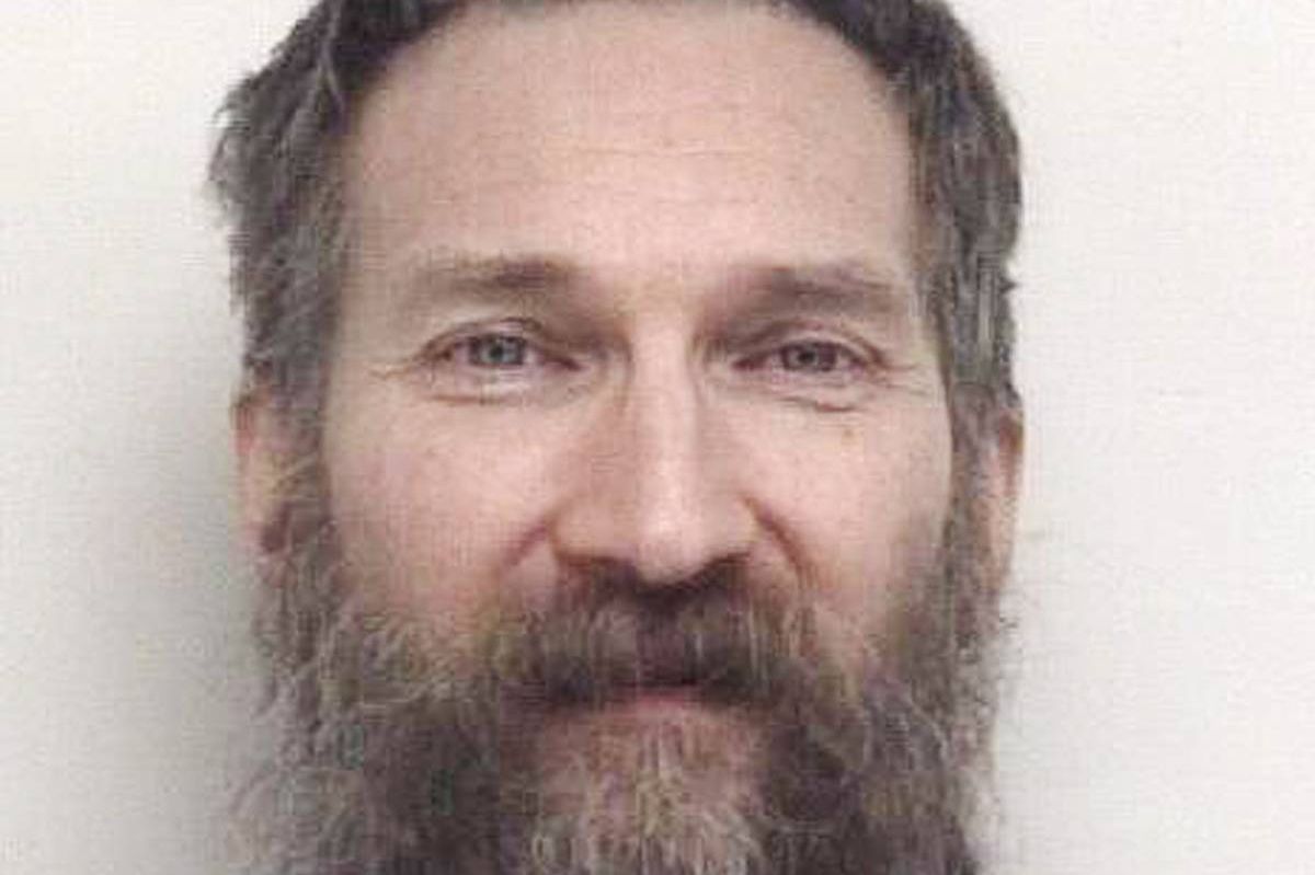 A close-up photograph depicts an individual with a full beard and mustache, exuding a contemplative expression. The subject's gaze is directed towards the camera, and the image is captured against a plain, light-colored background.