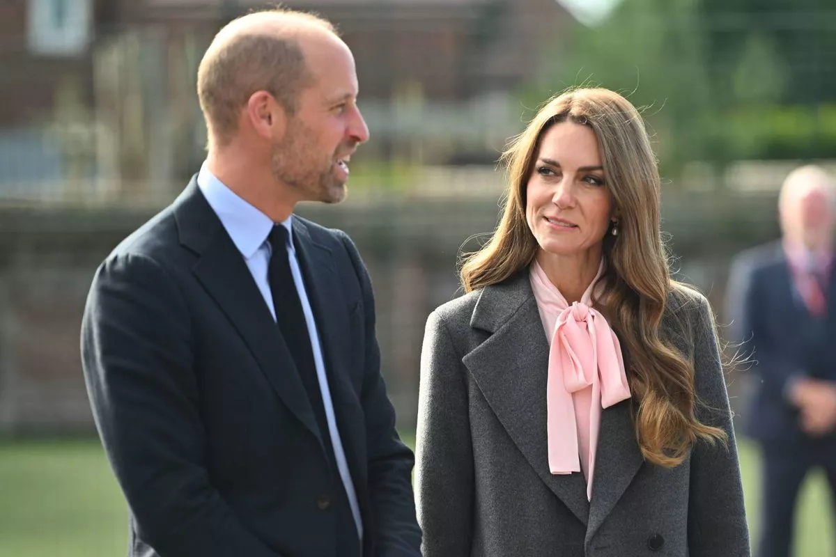 An individual in formal attire, consisting of a suit and tie, stands alongside a person wearing a blazer and a pink top, both engaged in conversation. The backdrop features a serene landscape with a body of water and greenery.