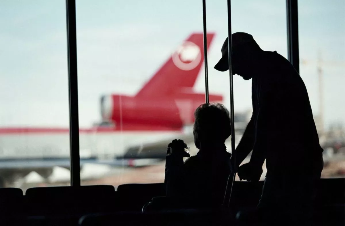 Silhouettes of individuals are visible in an airport setting, with a person standing and assisting another who is seated, while in the background, the tail of an airplane is prominently displayed.