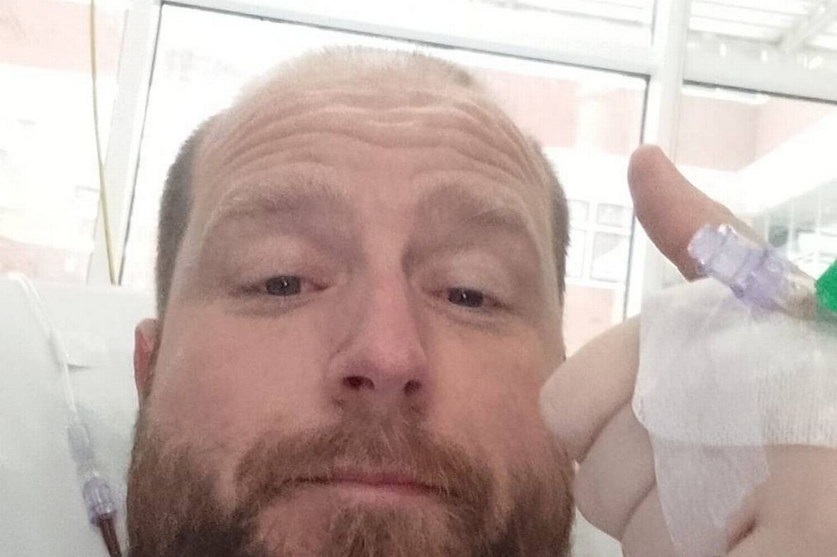 An individual with a beard and mustache is lying on a bed, holding a medical device with a bandage on his hand. The person's expression is one of contemplation, and the setting appears to be a hospital room with a window in the background.