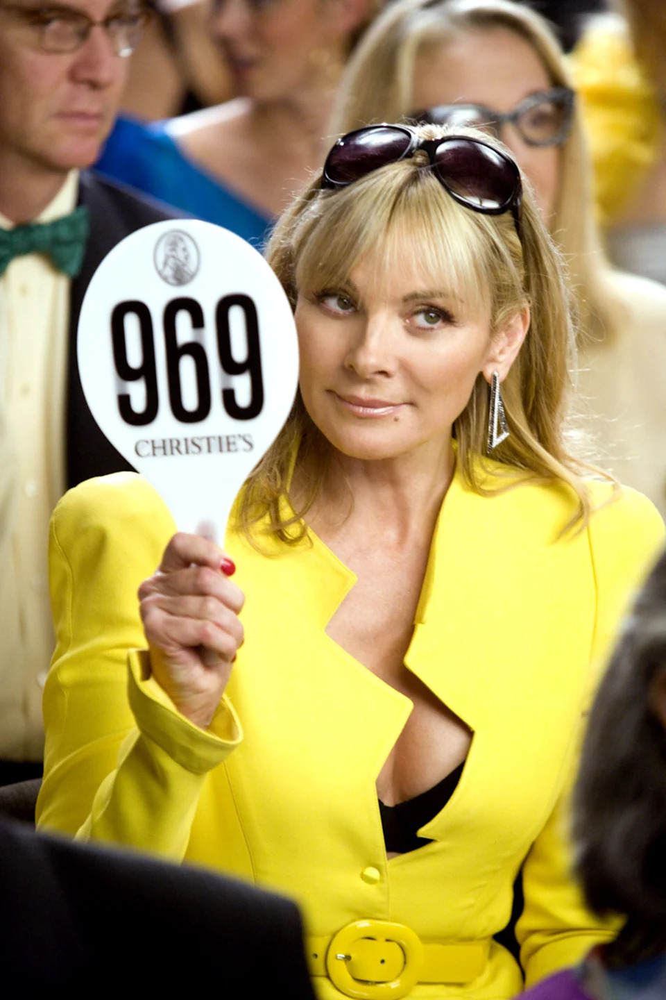 Person holding auction paddle with number 969, wearing stylish yellow blazer and sunglasses in hair, seated in an auction setting