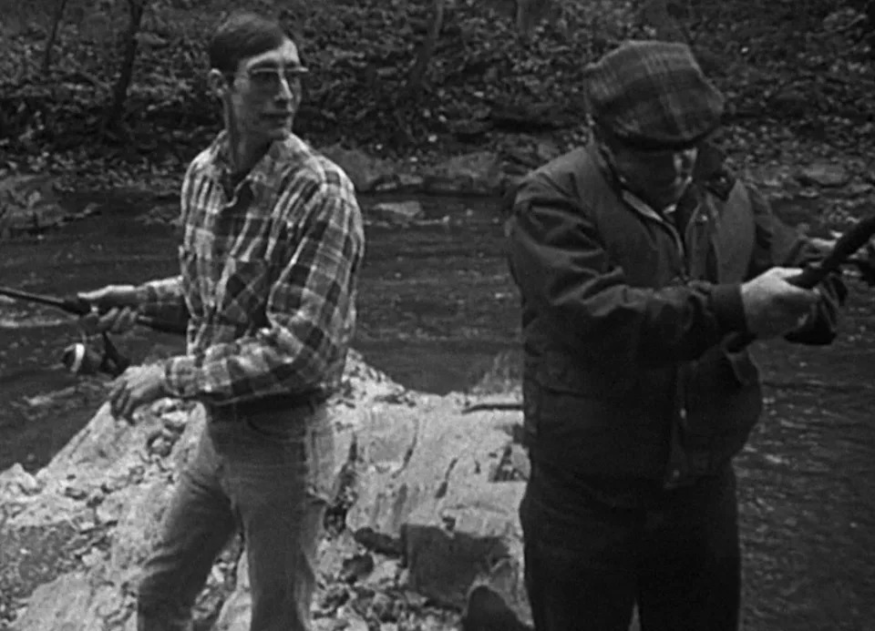 Two people stand on a rocky riverbank holding fishing rods, appearing focused on preparing their gear